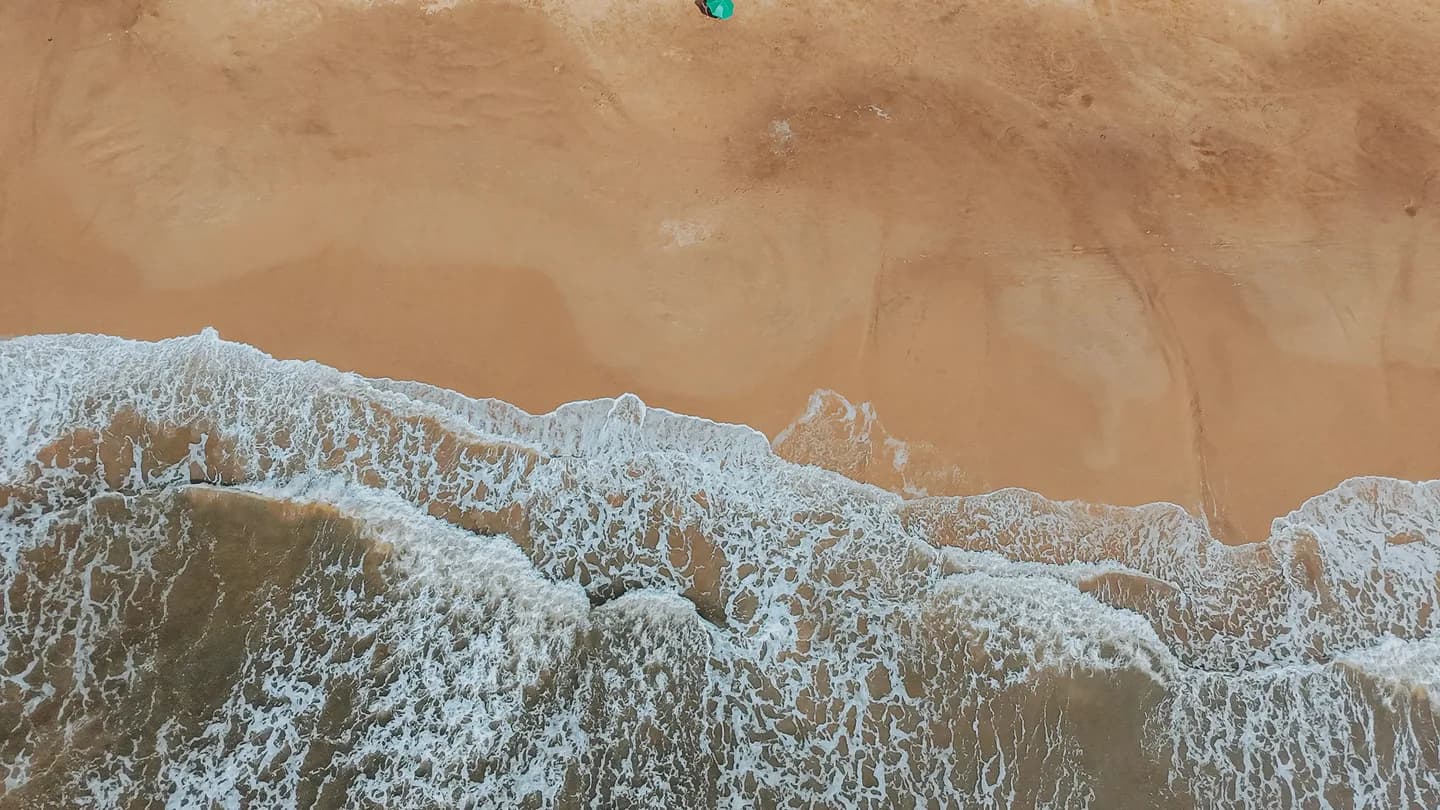 Straight-down aerial of surf meeting sand, water grading from white to deep blue. Bright sun, graphic and abstracted.