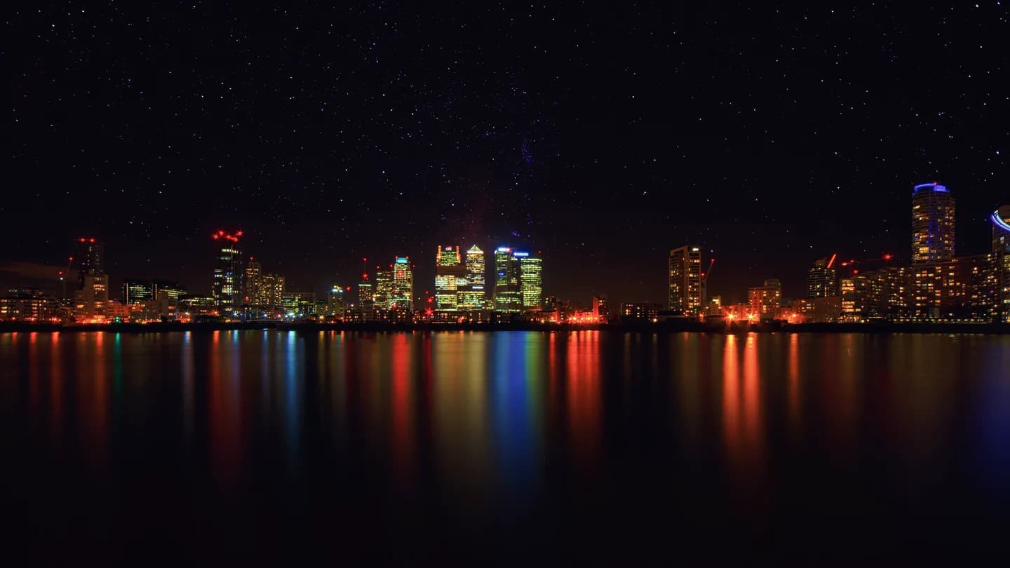 A city skyline after dark, windows and signage scattered across the towers. Long exposure, deep blue hour, wide and expansive.