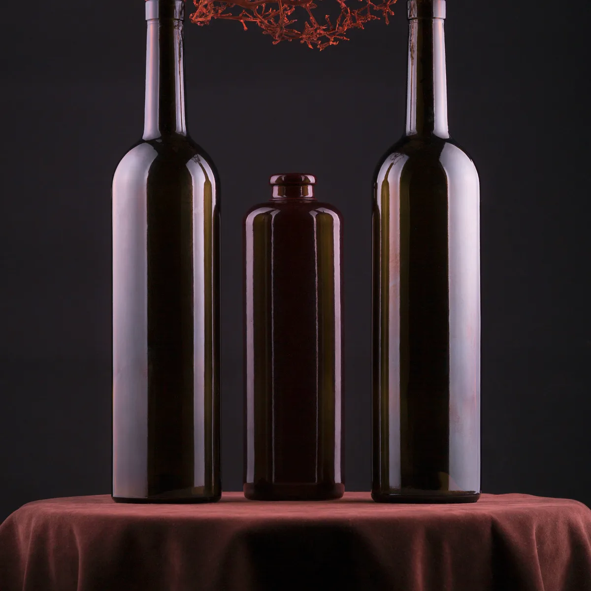 Three dark glass bottles grouped on draped cloth, lit hard from one side so only the shoulders catch. Deep shadow, muted plum and olive, a quiet still life.