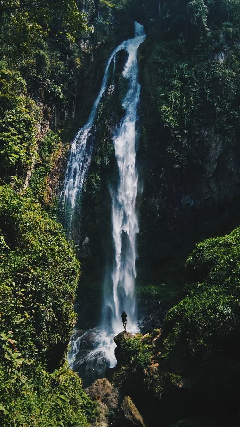 A thin waterfall dropping down a green rock face, spray softening the base. Long exposure smooths the water. Lush and cool.