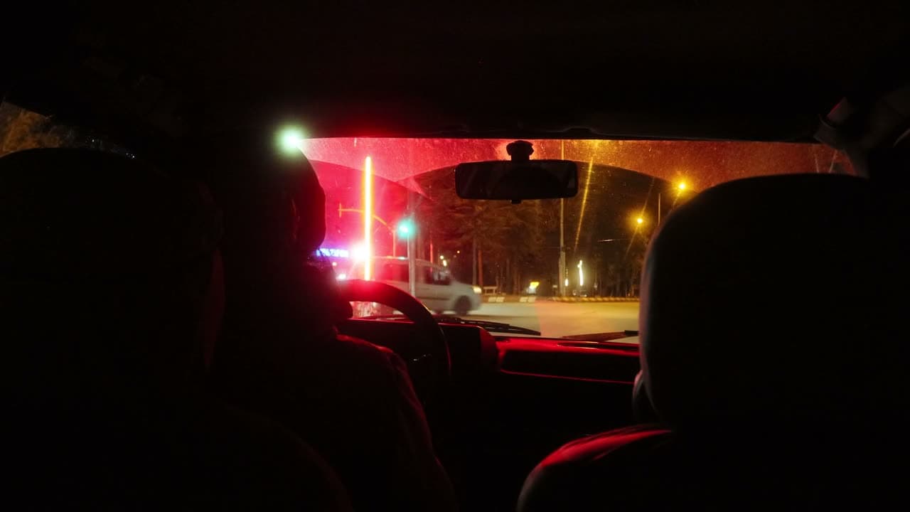 A car moves through city streets after dark, headlights and signage streaking past the windows. Tracking alongside, deep blacks, neon spill.