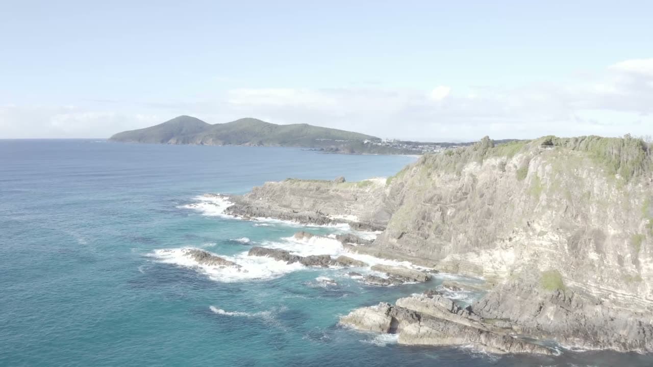 Drone glides along a cliff edge where surf meets rock, water turning turquoise in the shallows. Bright midday sun, wide and sweeping.