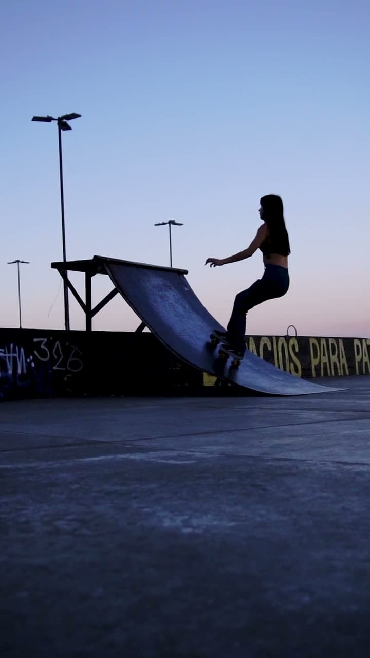 A skateboarder rolls through an empty concrete plaza, camera tracking alongside at knee height. Late afternoon sun, long shadows, loose documentary energy.