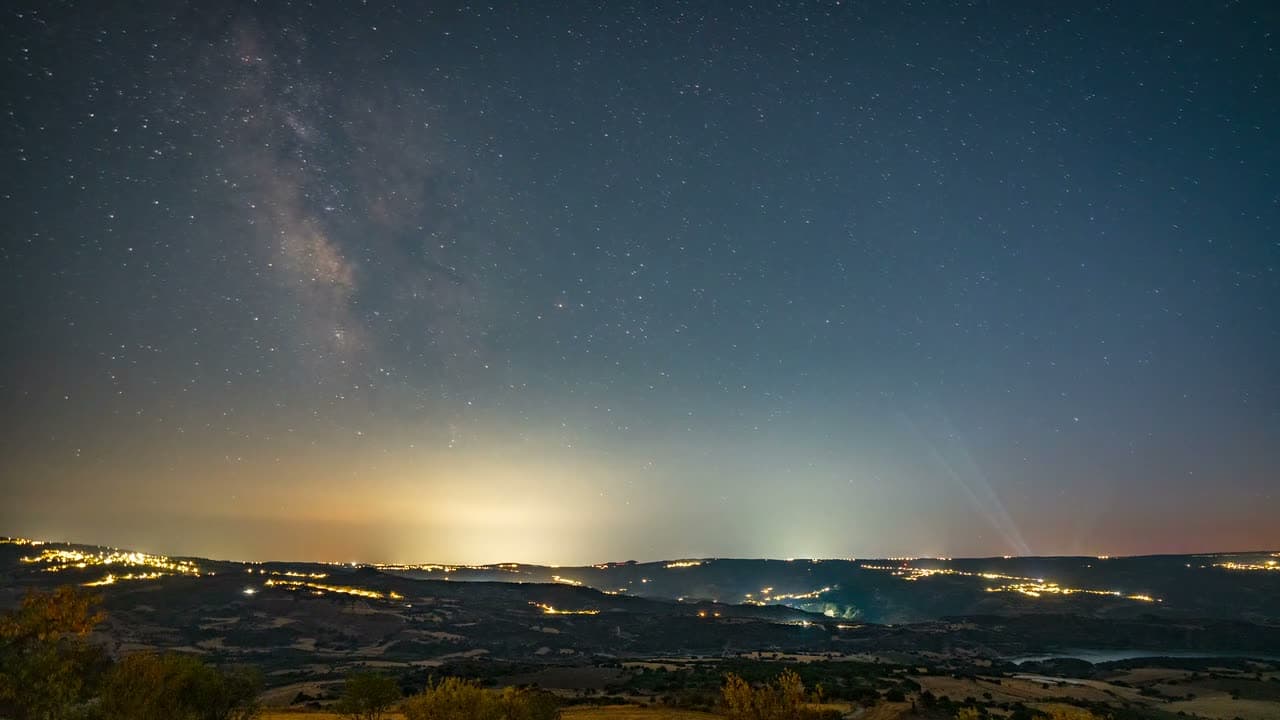 The Milky Way wheels slowly overhead in timelapse, horizon dark and low in frame. Dense starfield, cold blue, enormous and silent.